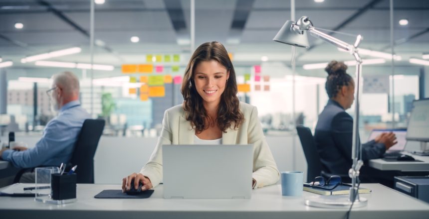 Young,Happy,Businesswoman,Using,Computer,In,Modern,Office,With,Colleagues.
