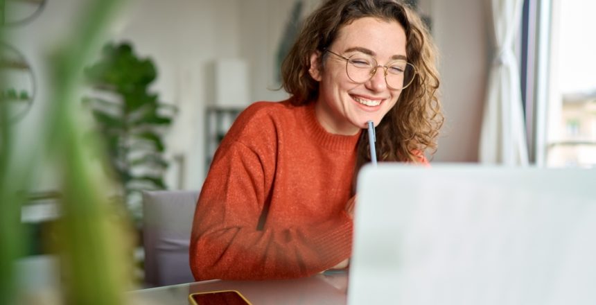 Happy,Young,Woman,Using,Laptop,Sitting,At,Desk,Writing,Notes